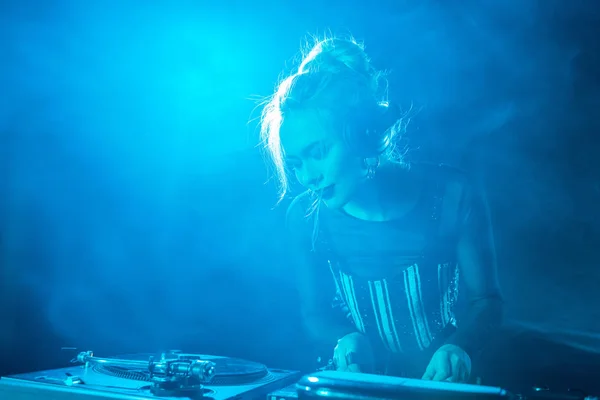 Glückliches blondes DJ-Mädchen, das Musik in Kopfhörern hört, während es DJ-Ausrüstung in einem Nachtclub mit Rauch benutzt — Stockfoto