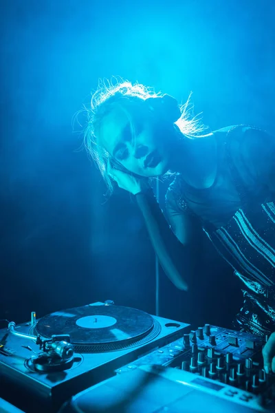 Beautiful dj girl listening music in headphones while standing with closed eyes in nightclub with smoke — Stock Photo