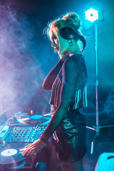 Hermosa mujer dj con cabello rubio escuchando música y sosteniendo auriculares en discoteca con humo - foto de stock