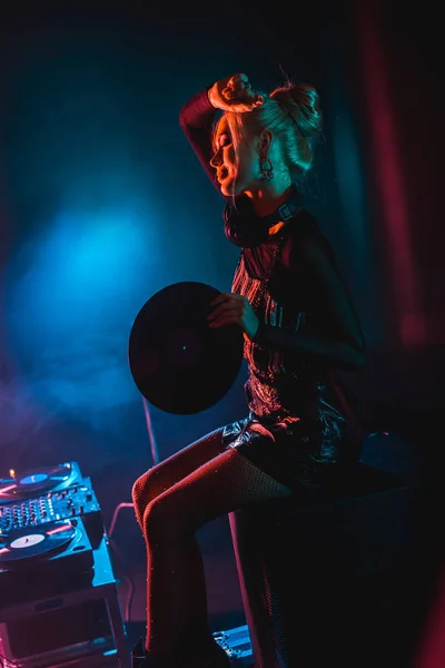 Cheerful blonde dj woman holding retro vinyl record in hand near dj equipment in nightclub with smoke — Stock Photo