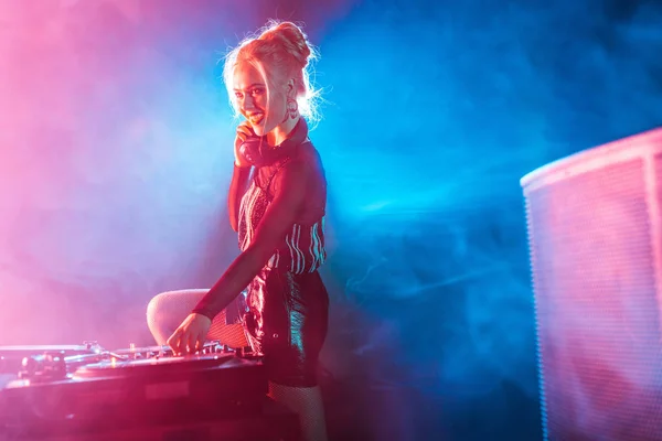 Gaie blonde dj femme dans les écouteurs en utilisant l'équipement dj dans la boîte de nuit avec de la fumée — Photo de stock