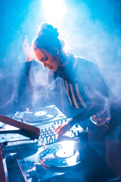 Concentrated dj woman with blonde hair standing in headphones and using dj equipment in nightclub with smoke — Stock Photo