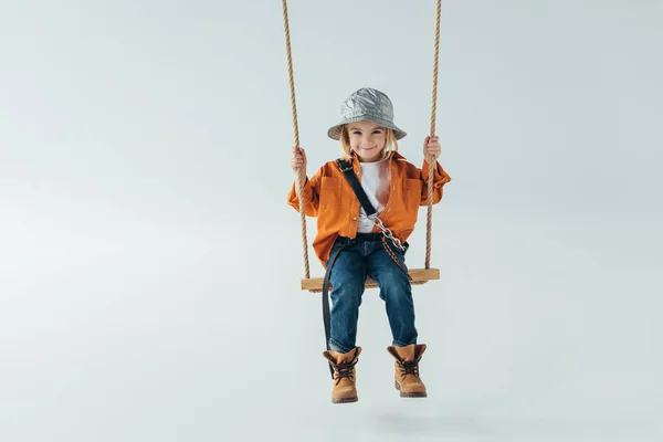 Cute kid in silver hat, jeans and orange shirt sitting on swing on grey background — Stock Photo