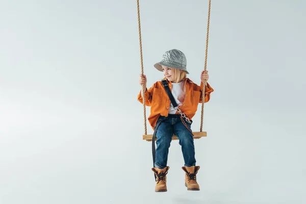 Lindo niño en jeans y camisa naranja sentado en el columpio y mirando hacia otro lado sobre fondo gris - foto de stock
