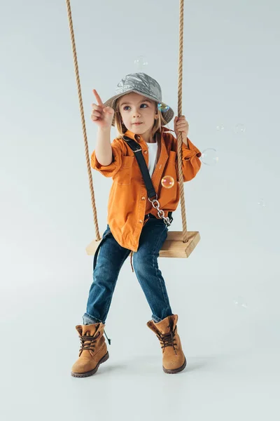 Cute kid in jeans and orange shirt sitting on swing and ponting with finger at soap bubbles — Stock Photo
