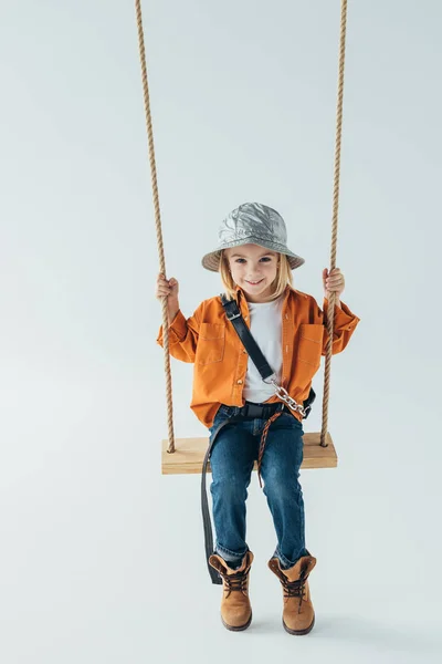 Enfant souriant en jeans et chemise orange assis sur balançoire sur fond gris — Photo de stock