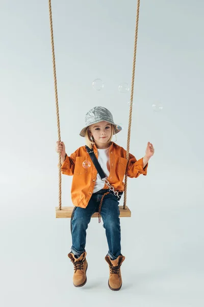 Cute kid in jeans and orange shirt sitting on swing and looking at soap bubbles — Stock Photo