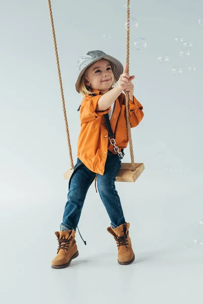 Smiling kid in jeans and orange shirt sitting on swing and ponting with finger at soap bubbles — Stock Photo