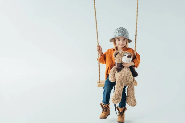 Cute kid in jeans and orange shirt sitting on swing and holding teddy bear — Stock Photo