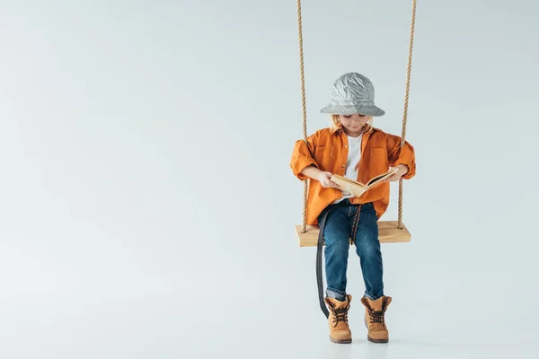 Cute kid in jeans and orange shirt sitting on swing and reading book on grey background — Stock Photo