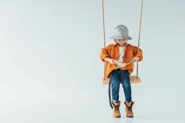 Garoto bonito em jeans e camisa laranja sentado no swing e livro de leitura em fundo cinza — Fotografia de Stock