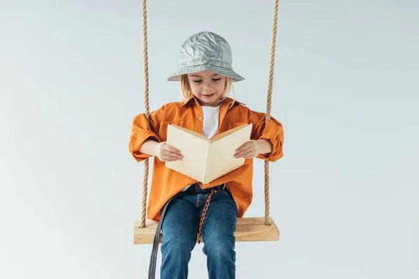 Adorable kid in jeans and orange shirt sitting on swing and reading book isolated on grey — Stock Photo