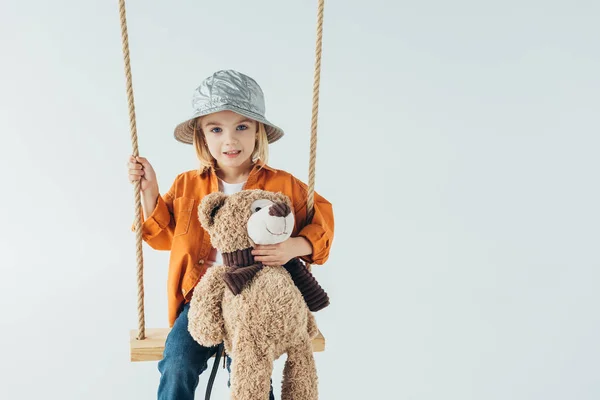 Cute kid sitting on swing and holding teddy bear isolated on grey — Stock Photo