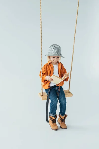 Adorable enfant en jeans et chemise orange assis sur balançoire et livre de lecture — Photo de stock