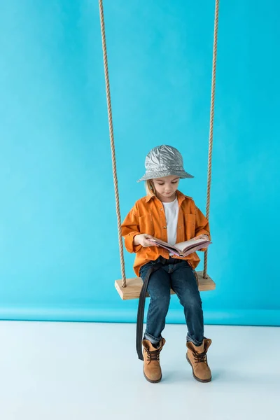 Adorable enfant en jeans et chemise orange assis sur balançoire et livre de lecture sur fond bleu — Photo de stock