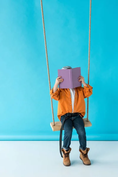 Cute kid in jeans and orange shirt sitting on swing and reading book on blue background — Stock Photo