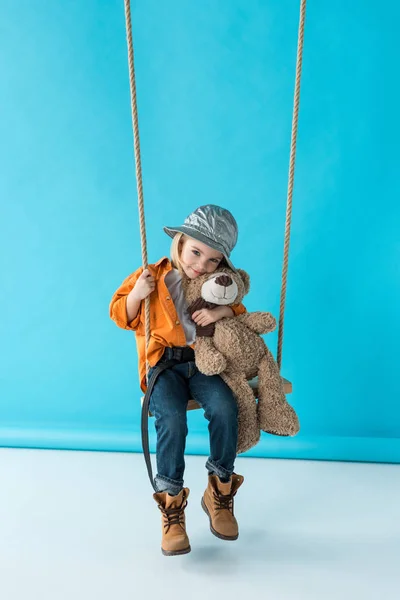 Lindo niño sentado en swing y abrazando osito de peluche sobre fondo azul - foto de stock