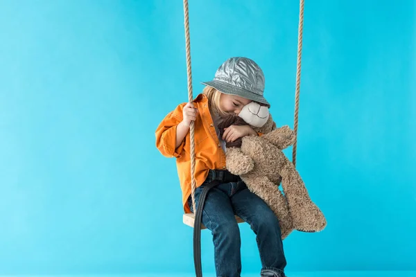 Милый ребенок сидит на качелях и обнимает плюшевого медведя изолированы на синий — стоковое фото