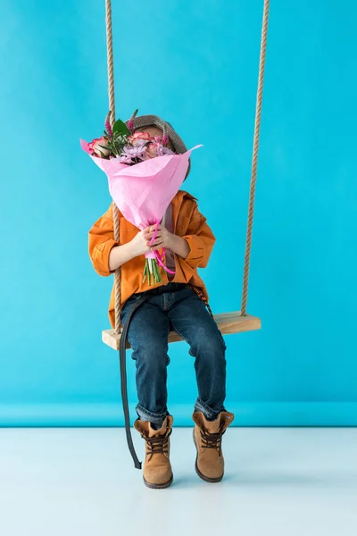 Kind sitzt auf Schaukel und hält Blumenstrauß auf blauem Hintergrund — Stockfoto