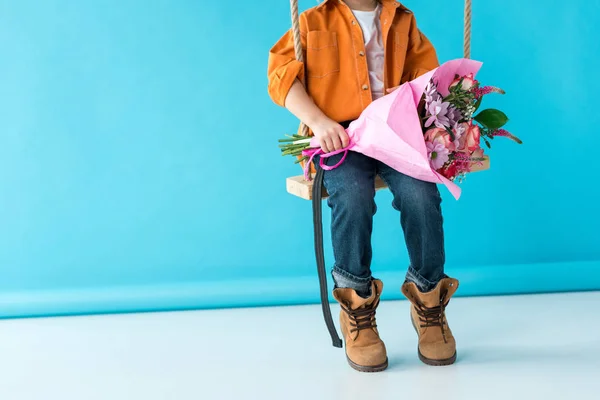 Cropped view of kid sitting on swing and holding bouquet on blue background — Stock Photo