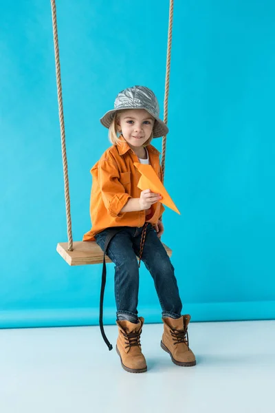 Mignon enfant assis sur balançoire et tenant avion en papier sur fond bleu — Photo de stock