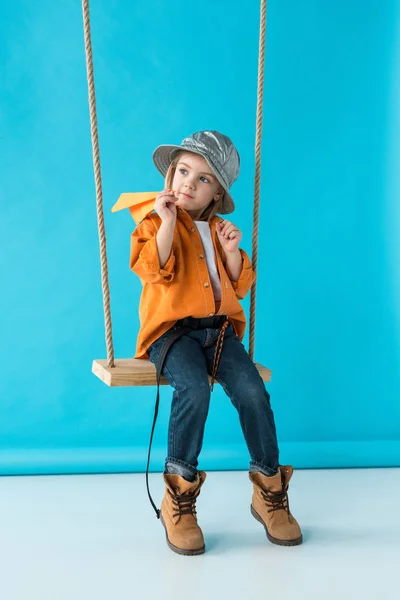 Cute kid sitting on swing, holding paper plane and looking away on blue background — Stock Photo