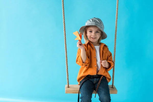 Niedliches Kind sitzt auf Schaukel, hält Papierflieger in der Hand und blickt isoliert in die Kamera auf blauem Grund — Stockfoto