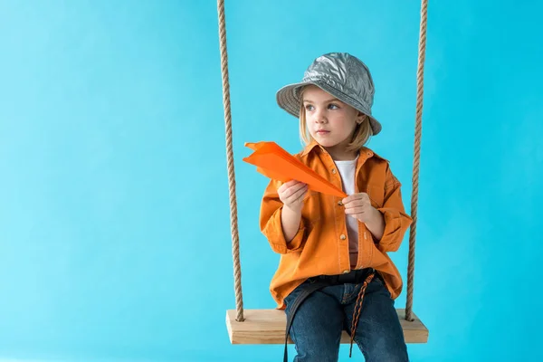 Cute child sitting on swing, holding paper plane and looking away isolated on blue — Stock Photo