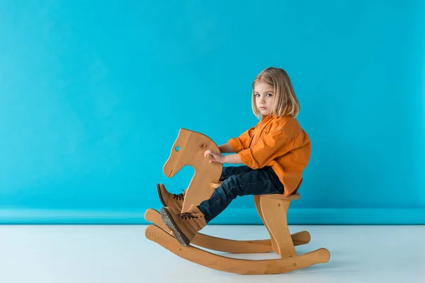 Serious and cute kid sitting on rocking horse and looking at camera on blue background — Stock Photo