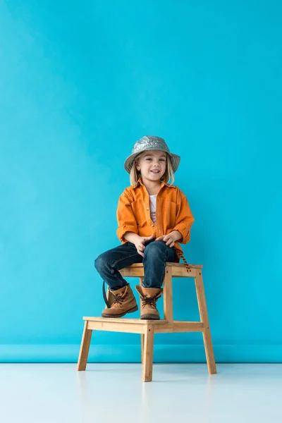 Niño sonriente en jeans y camisa naranja sentado en las escaleras y mirando a la cámara - foto de stock