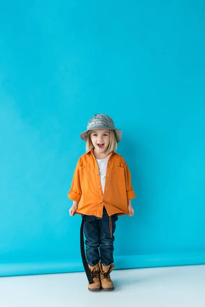 Surpised kid in silver hat, jeans and orange shirt looking away on blue background — Stock Photo