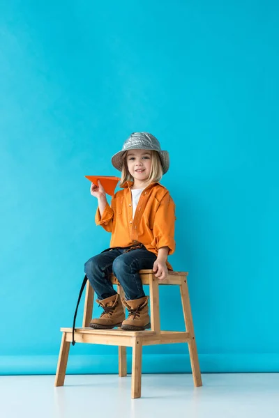 Kid in jeans and orange shirt sitting on stairs and holding orange plane on blue background — Stock Photo