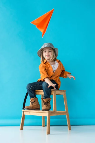 Niño en jeans y camisa naranja sentado en las escaleras y jugando con avión de papel - foto de stock