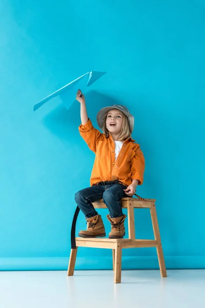 Bambino in jeans e camicia arancione seduto sulle scale e giocare con aereo di carta blu — Foto stock