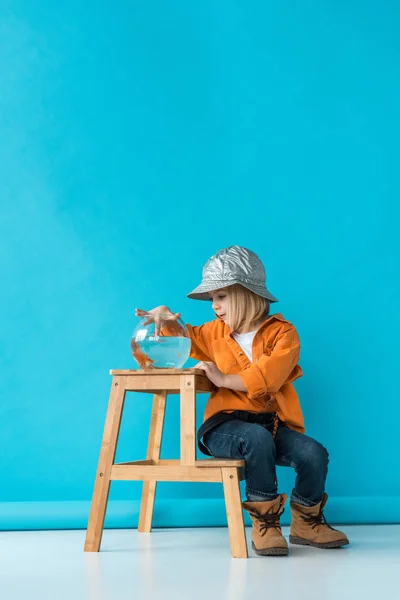 Niño en jeans y camisa naranja sentado en las escaleras y jugando con peces de colores - foto de stock