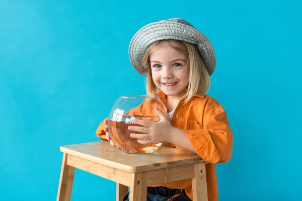 Enfant en chapeau argenté et chemise orange tenant aquarium avec poisson rouge isolé sur bleu — Photo de stock
