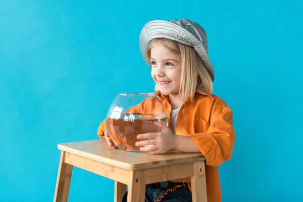 Bambino sorridente in cappello d'argento e camicia arancione che tiene acquario con pesci rossi isolati su blu — Foto stock
