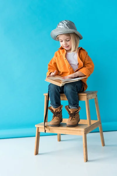 Kid in jeans and orange shirt sitting on stairs and reading book on blue background — Stock Photo