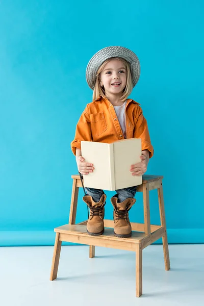 Niño con sombrero plateado y camisa naranja sentado en las escaleras y sosteniendo el libro sobre fondo azul - foto de stock
