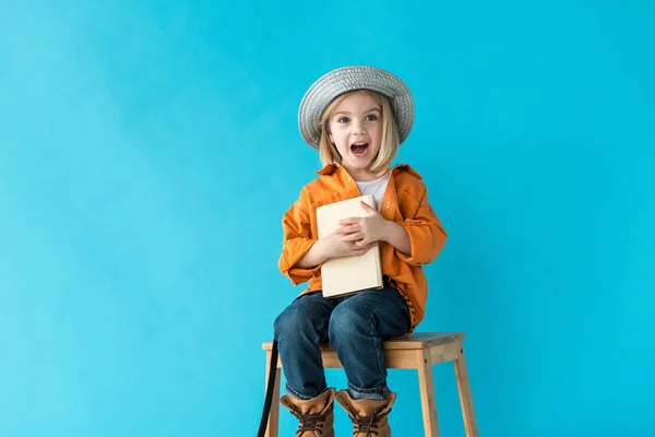 Niño sorprendido en jeans y camisa naranja sentado en las escaleras y sosteniendo libro aislado en azul - foto de stock