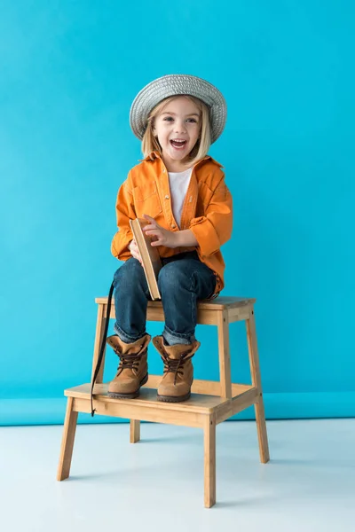 Enfant souriant en jeans et chemise orange assis sur les escaliers et tenant livre — Photo de stock