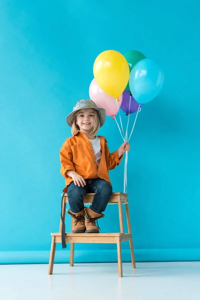 Lindo niño en jeans y camisa naranja sentado en escaleras y sosteniendo globos - foto de stock