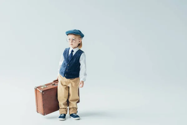 Дитина в ретро жилеті і кепці тримає валізу на сірому фоні — стокове фото