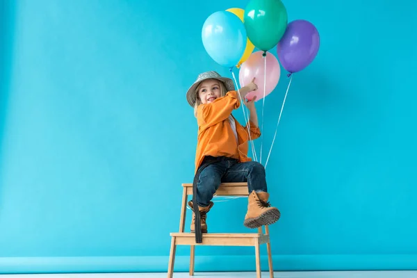 Lindo niño en jeans y camisa naranja sentado en las escaleras y apuntando con el dedo a los globos - foto de stock