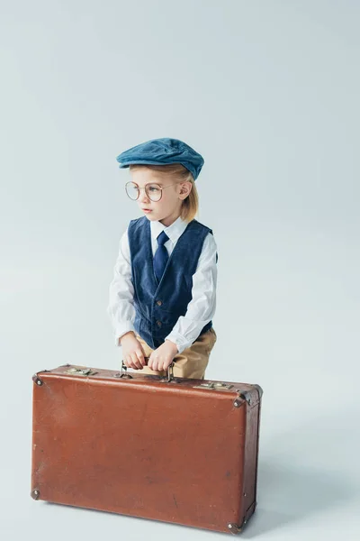 Вдумчивый ребенок в ретро жилете и кепке с чемоданом на сером фоне — стоковое фото