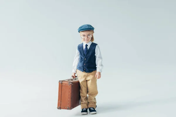Lächelndes Kind in Retro-Weste und Mütze mit Koffer auf grauem Hintergrund — Stockfoto