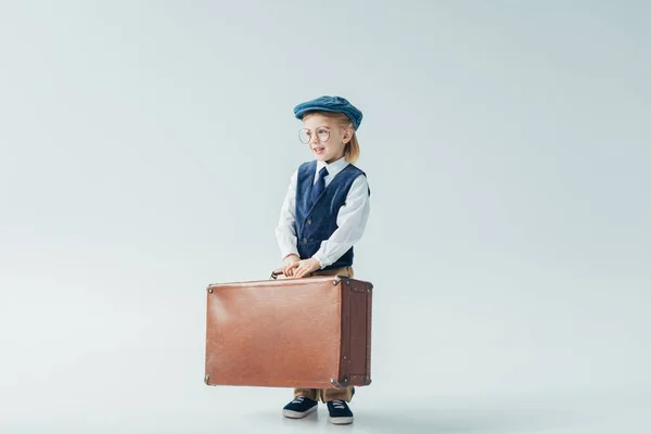 Усміхнена дитина в ретро жилеті і кепці тримає валізу і дивиться на сірий фон — стокове фото