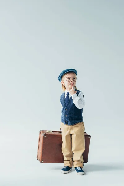 Мечтательный ребенок в ретро жилете и кепке держа чемодан и глядя в сторону на сером фоне — стоковое фото