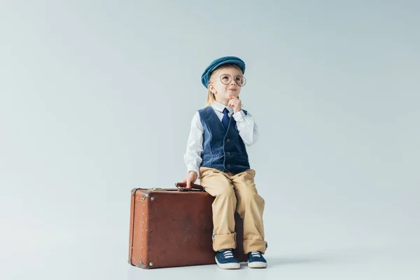 Cher enfant en gilet rétro et casquette assis sur la valise sur fond gris — Photo de stock