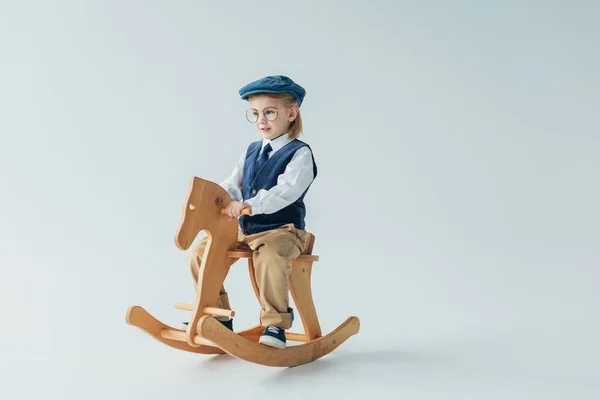 Ребенок в ретро жилете и кепке катается на качающейся лошади и смотрит в сторону — стоковое фото